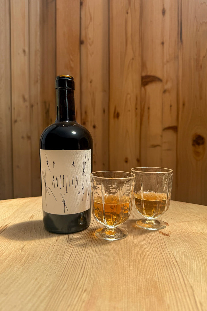 A bottle of Broc Cellars 2020 Angelica with wine glasses sits on a light wooden table, each glass filled with golden-brown wine. Behind, warm-toned vertical wood panels evoke the look of caramelized figs.