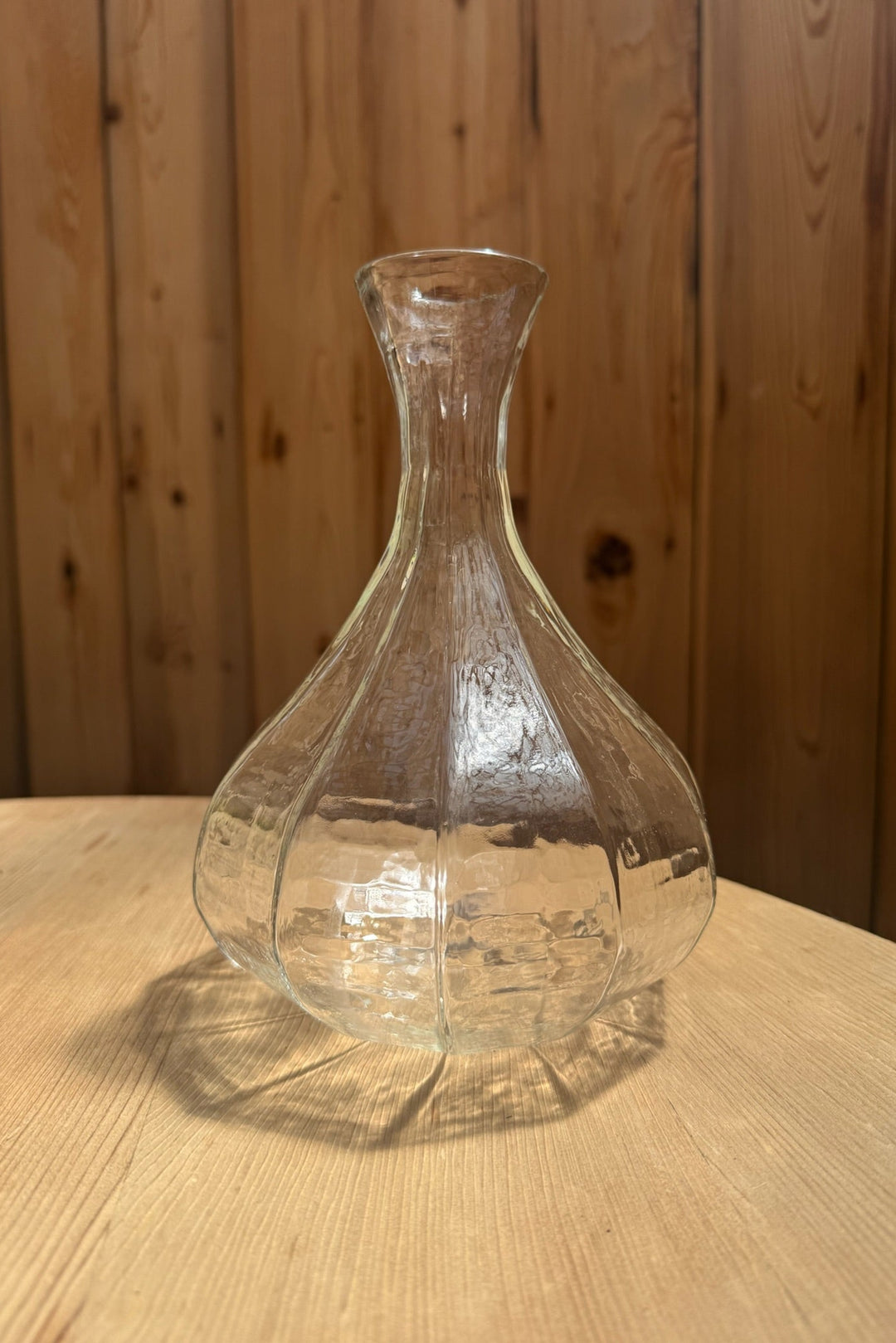 A clear Broc Cellars Decanter with a bulbous, ribbed base and narrow neck sits on a light wooden surface against vertical wooden planks—an elegant piece of handmade glassware.