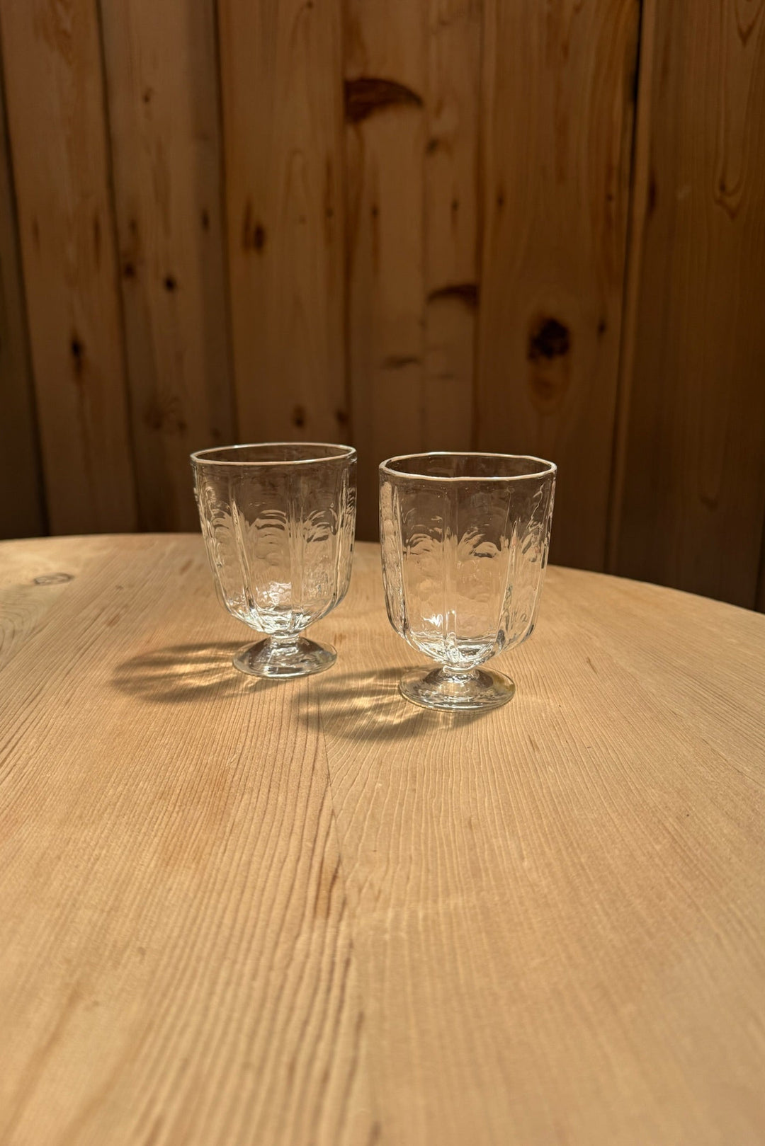 Two small Broc Glass goblets by Broc Cellars, each clear and handblown with etched designs, rest on a light wood table. A vertical plank wooden wall forms the background as the glasses cast delicate shadows below.