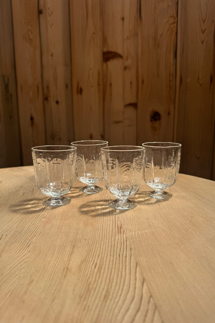 Four patterned Broc Glass wine glasses by Broc Cellars sit on a light wooden table, their handblown design casting soft shadows, with vertical wooden panels in the background.