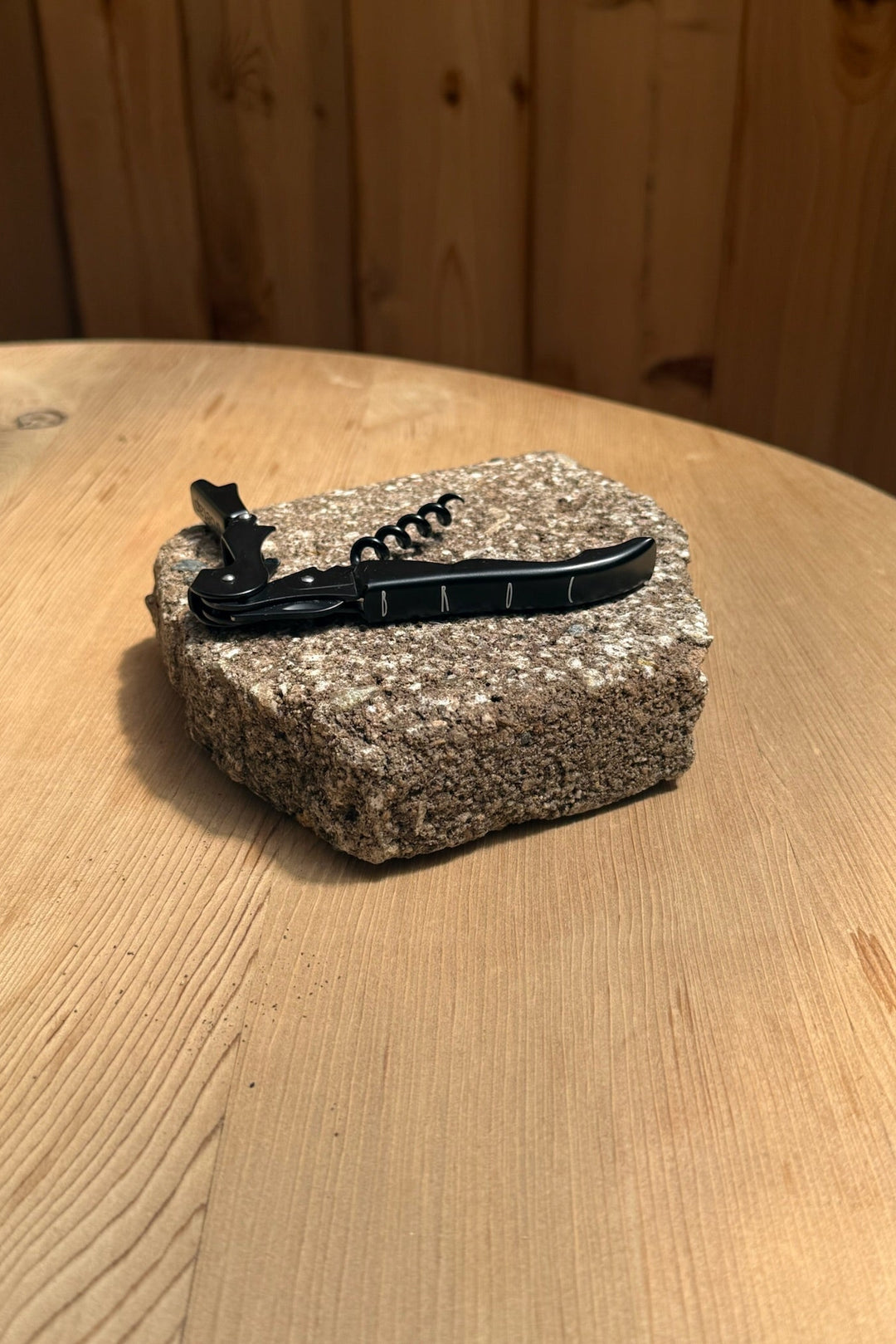 The Broc Cellars Black Wine Key and bottle opener sits atop a rectangular, rough stone on a wooden table with a wood-paneled wall behind it.