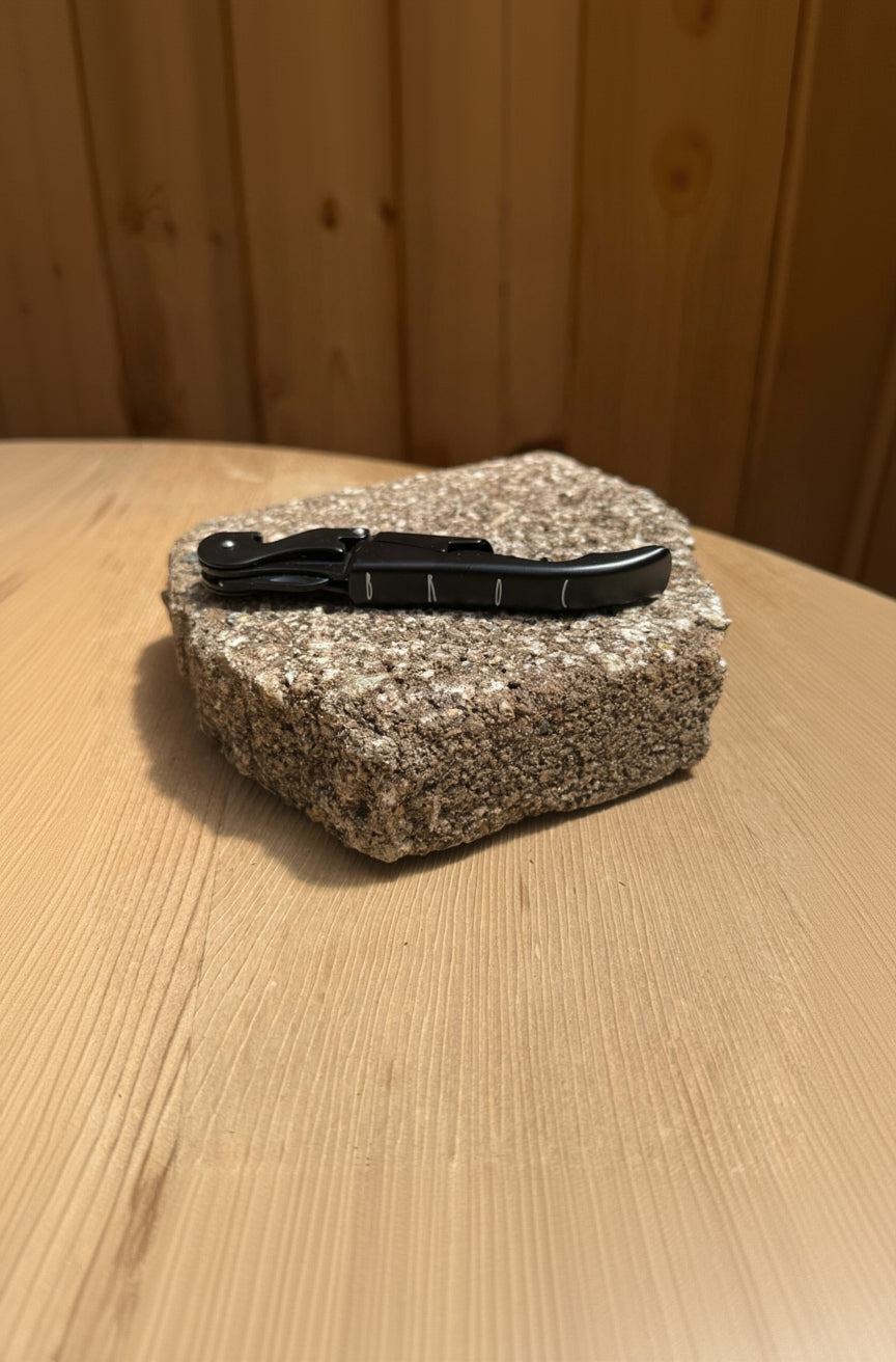 A Broc Cellars Black Wine Key, sleek and matte black, rests beside a folded pocket knife on granite atop a light wooden surface with a wood-paneled background, adding modern flair.