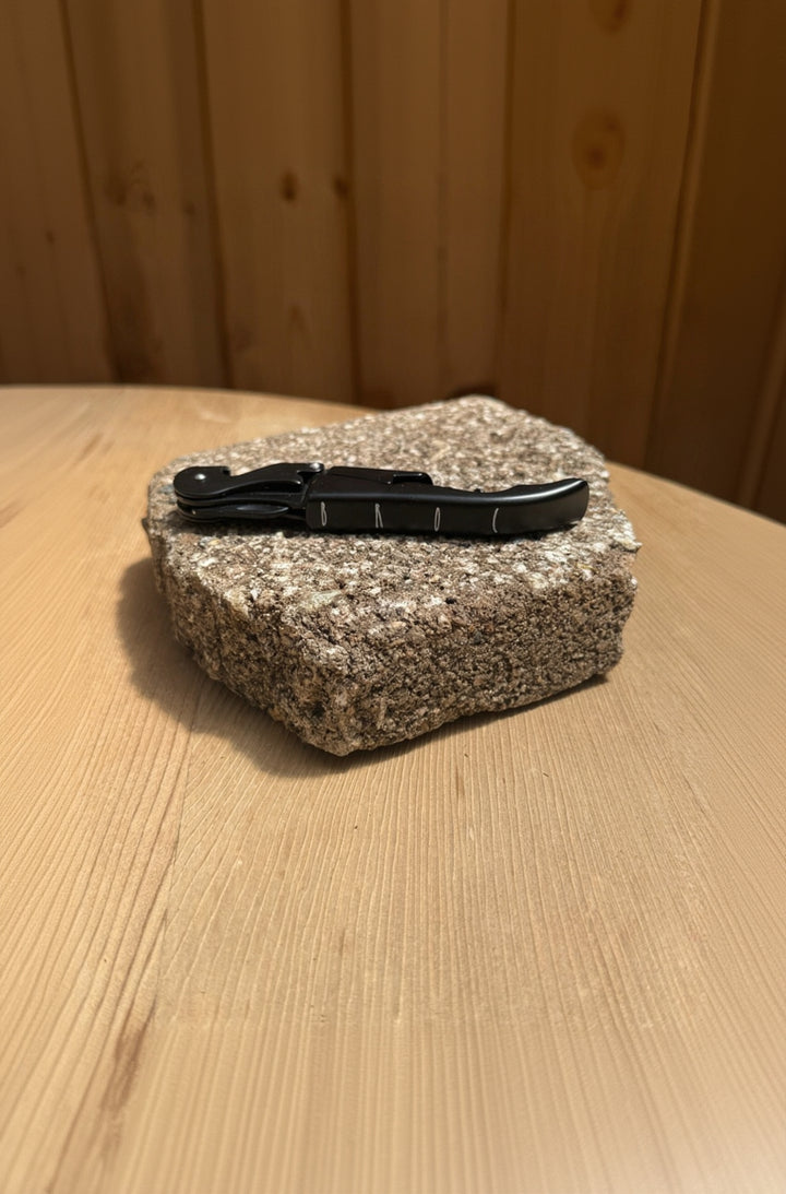 A Broc Cellars Black Wine Key, sleek and matte black, rests beside a folded pocket knife on granite atop a light wooden surface with a wood-paneled background, adding modern flair.