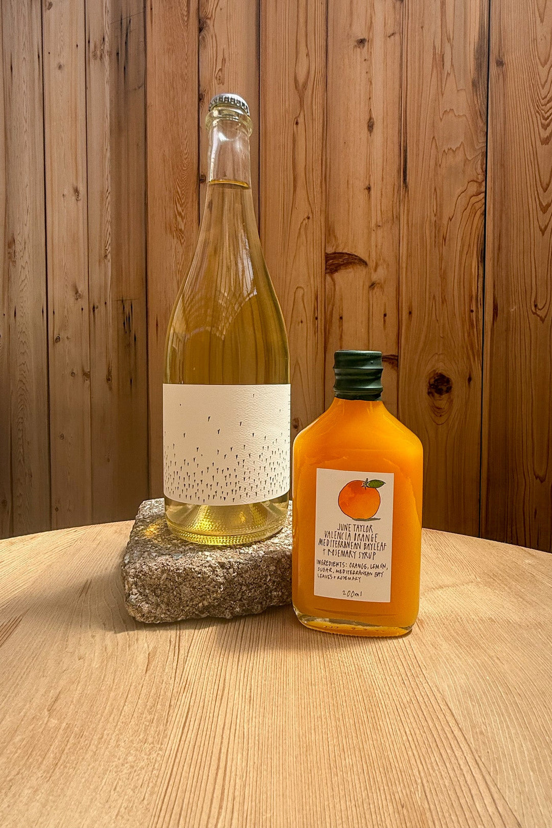 A clear Broc Cellars Spritz Pack bottle with a blank cream label and yellow beverage stands on a stone beside a smaller June Taylor Seasonal Fruit Syrup bottle. Both are on a wooden table with wooden paneling in the background.