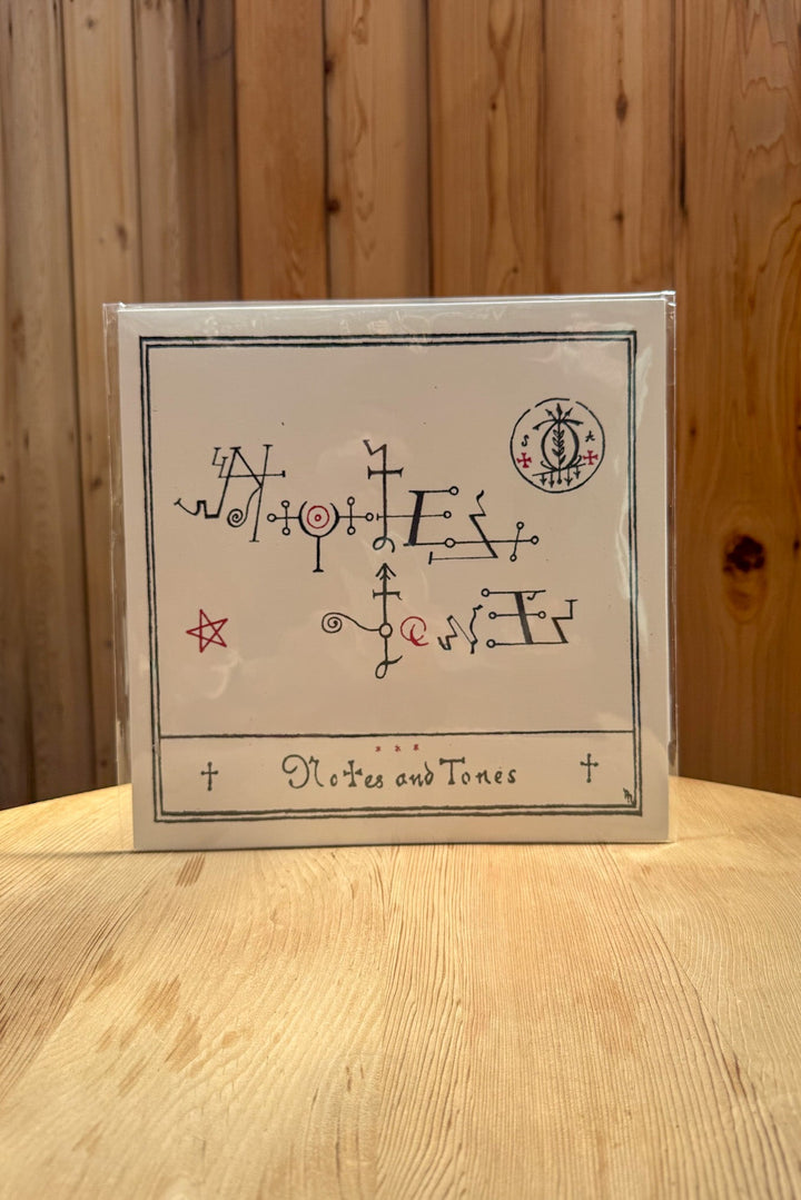A square card for Notes &amp; Tones V.1 - The Album by Broc Cellars features black and red abstract symbols framed by a thin border. The card sits on a light wood table against a wood-paneled wall, evoking cosmic jazz.
