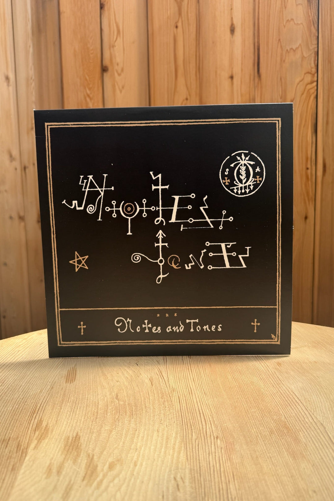 A black square box labeled “Notes & Tones V.2 - The Album” by Broc Cellars, adorned with gold-outlined symbols and sigils, sits on a light wooden surface against vertical wood panels.