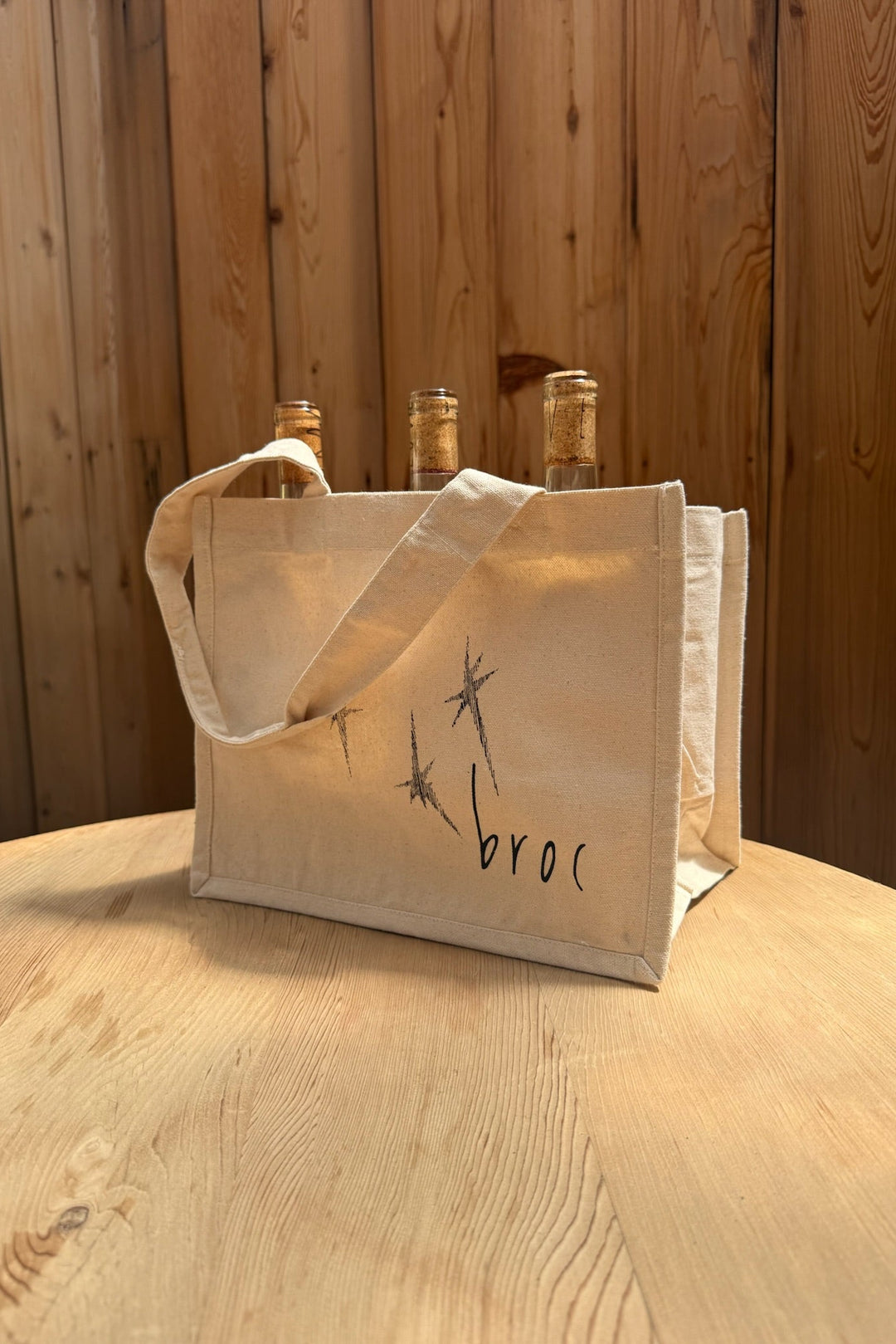 The Broc Bottle Tote by Broc Cellars is a beige canvas bag with two handles, bottle compartments for three bottles, and minimalist “broc” branding with line art. It sits on a wooden table against wooden paneling.