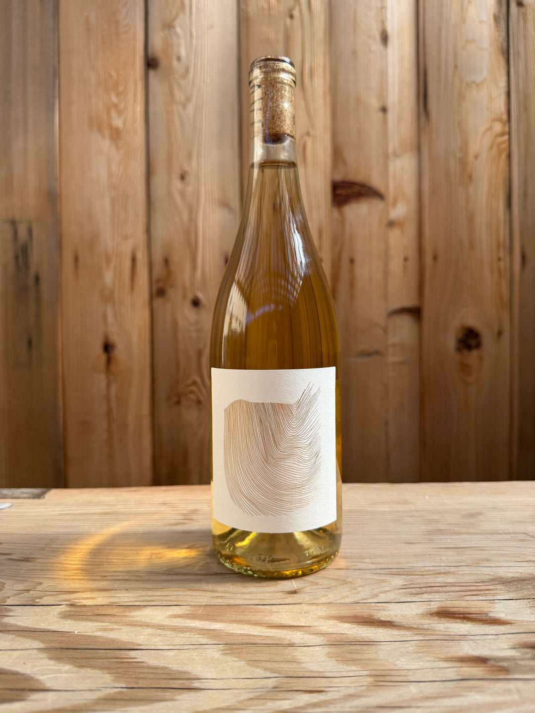 A clear glass wine bottle with a cork sits on a wooden surface. The bottle, labeled “Erica Tanov x Broc” from Broc Cellars, features a minimalistic white label adorned with a simple, abstract brushstroke design. Vertical wood panels create the background.
