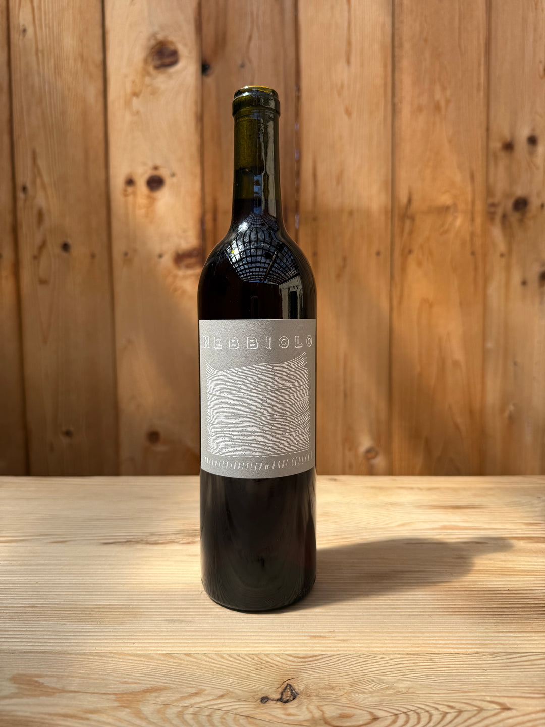 A single dark wine bottle with a minimal, light-colored label reading “2025 Nebbiolo” sits on a light wooden table in front of a vertical wooden plank wall, sunlight casting a shadow across the surface. This exclusive release from Broc Cellars is available to Wine Club members only.