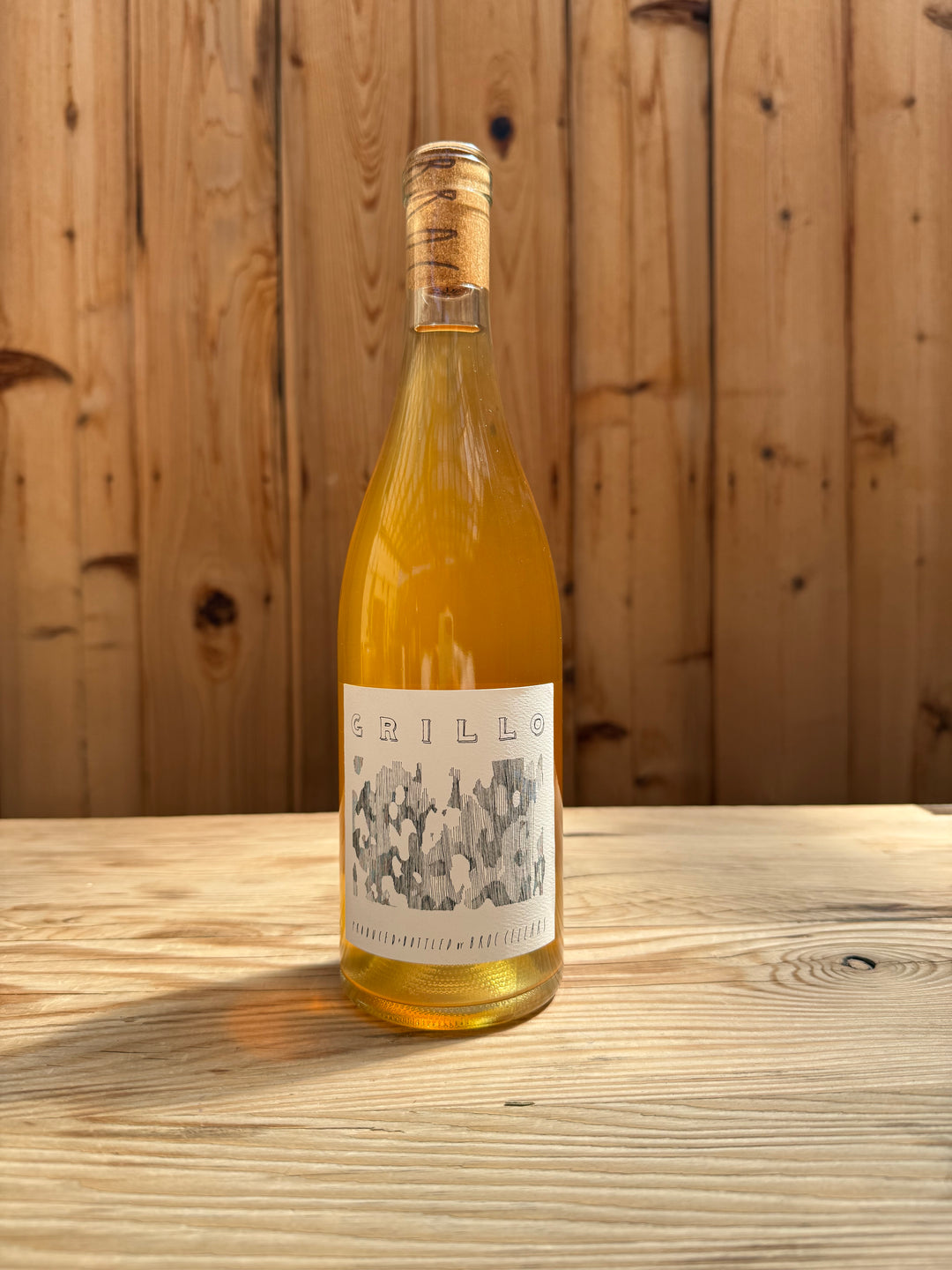 A bottle of 2025 Grillo from Broc Cellars, marked “Wine Club Only,” with a cork and illustrated label rests on a wooden surface, set against a wooden wall in the background.