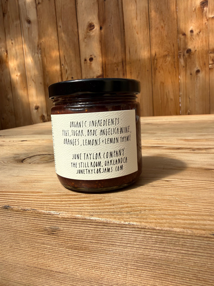 A glass jar of Broc Cellars’ June Taylor Mission Fig + Angelica Jam sits on a wooden surface with a wood fence in the background. The label highlights its organic ingredients—figs, sugar, Bosc and Seckel pears, oranges, lemons, and lemon thyme—emphasizing the artisanal nature of this fig preserve.