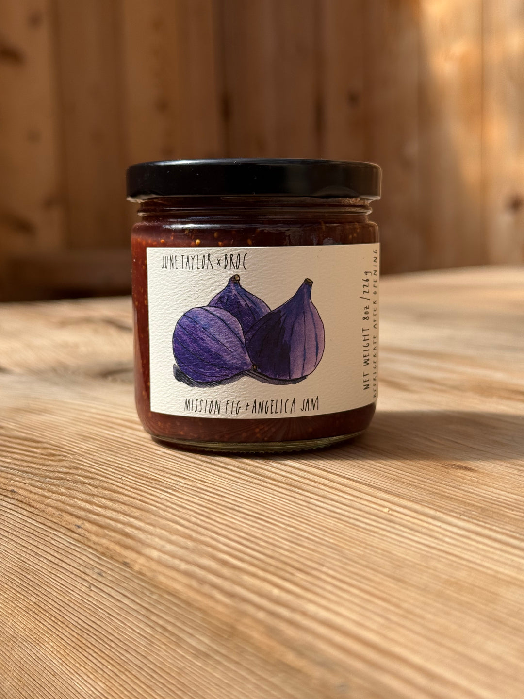 A jar of Broc Cellars June Taylor Mission Fig + Angelica Jam rests on a wooden surface. The label features an illustration of two figs along with product details in black text. Softly blurred wood paneling forms the background.