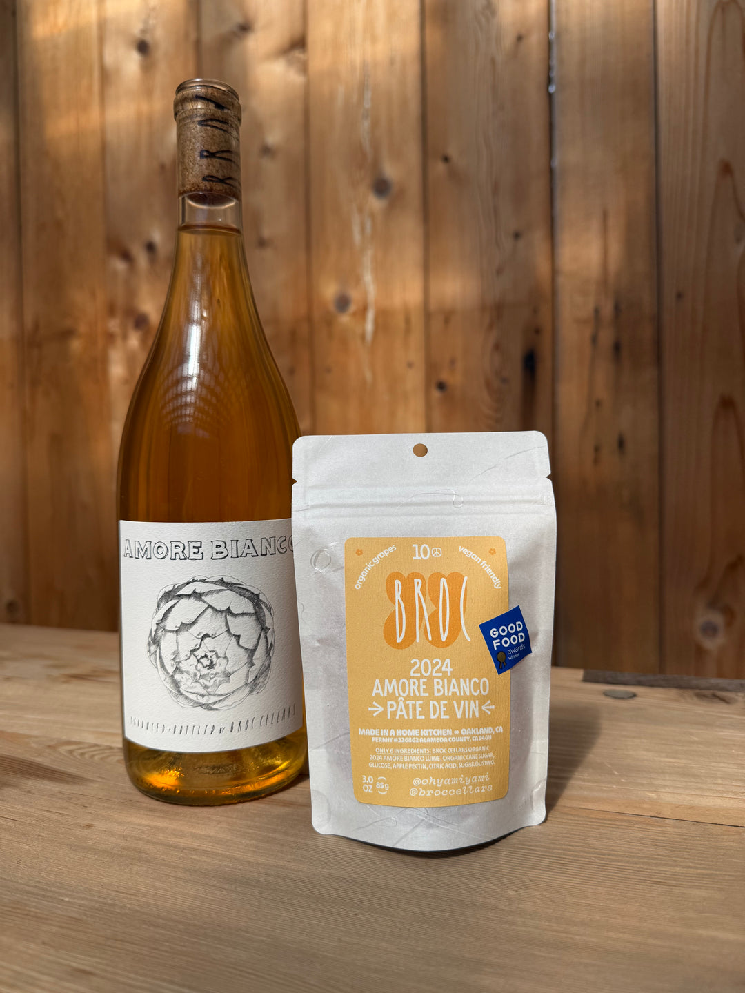 A bottle of Broc Cellars Amore Bianco + Yami Yami Pâte de Vin stands on a wooden surface next to a beige pouch labeled 2024 Amore Bianco Pâte de Vin, a vegan-friendly candy, set against a wooden wall background.