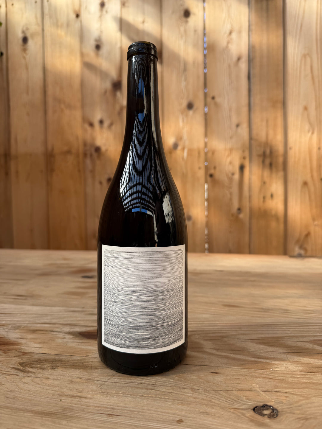 A dark glass bottle of Broc Cellars 2018 Counoise, featuring a blank, textured white label, sits on a wooden table. The background showcases wood paneling with visible knots and vertical seams, evoking the rustic charm befitting this medium-bodied red made from Eaglepoint Ranch grapes and aged in neutral French oak.
