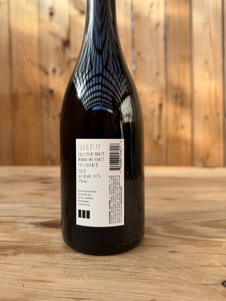 A dark glass wine bottle of Broc Cellars 2018 Counoise rests on a wooden surface with its label facing the camera. The label displays details about the wine, including notes of neutral French oak aging and a mention of Counoise Eaglepoint Ranch, along with a barcode and information about the producer. Wooden paneling forms the background.