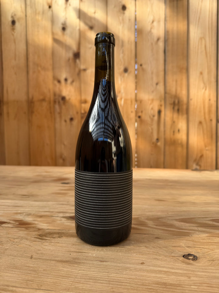 A dark glass bottle of 2019 Broc Valdiguié from Broc Cellars, distinguished by its black label with horizontal silver lines, sits on a wooden surface. The Library Release vintage wine is displayed against a background of vertical wooden planks illuminated by warm light and shadow.