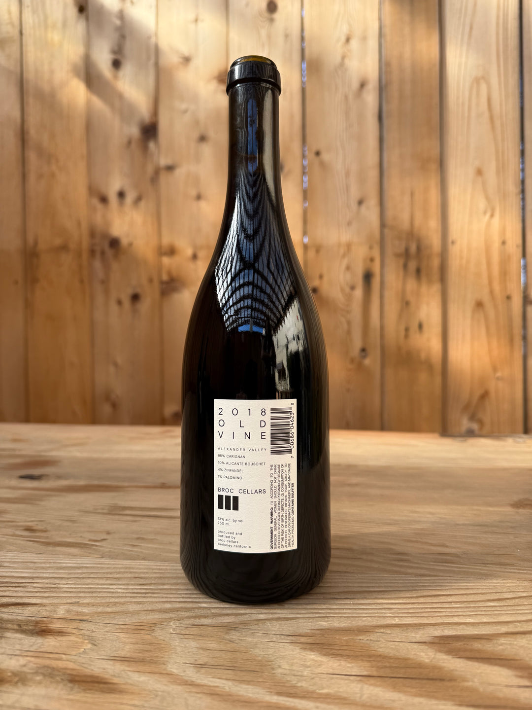 A dark glass wine bottle labeled “2018 OLD VINE CARIGNAN” from Broc Cellars, crafted from ancient vines, stands on a light wooden surface with a wooden plank wall in the background. The label displays details about the wine and a barcode.