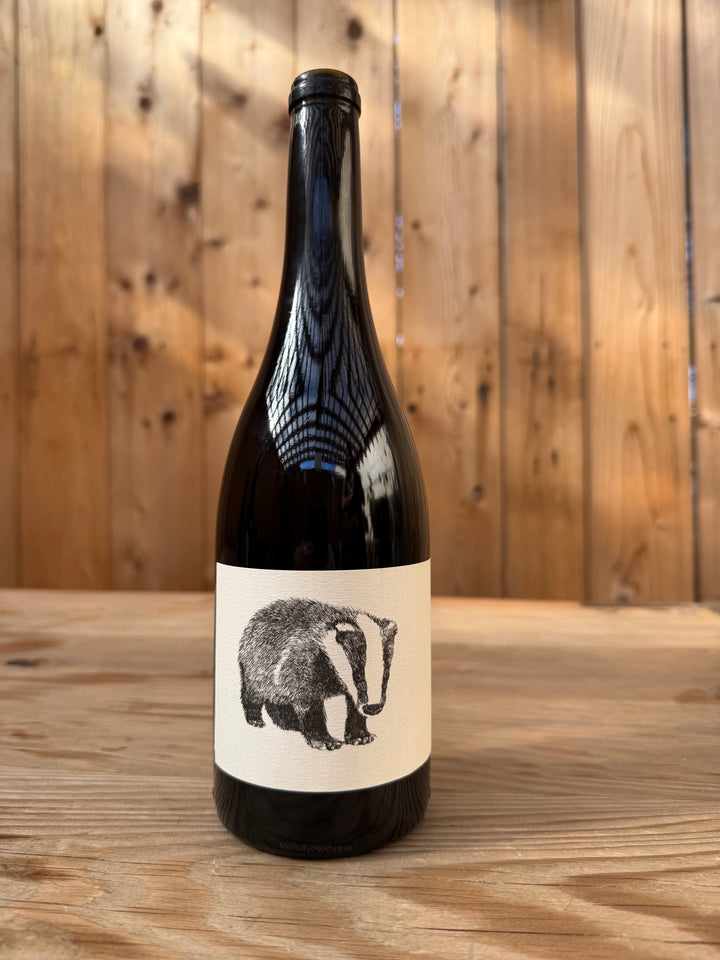 A dark glass bottle of Broc Cellars 2020 Nero d’Avola, featuring a white label with a black and white badger illustration, stands on a wooden surface against a wood plank background. This California wine is crafted from Nero d’Avola grapes sourced from the renowned Fox Hill Vineyard.