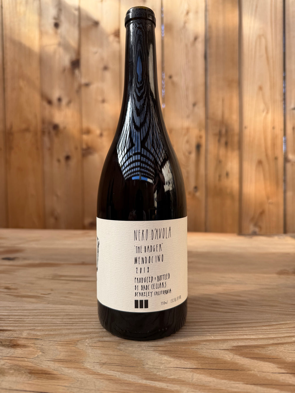 A bottle of 2018 NERO D’ AVOLA from Broc Cellars sits on a wooden surface, framed by a wooden wall in the background. The label displays its origin and vintage, with the bottle catching glints of overhead lights.