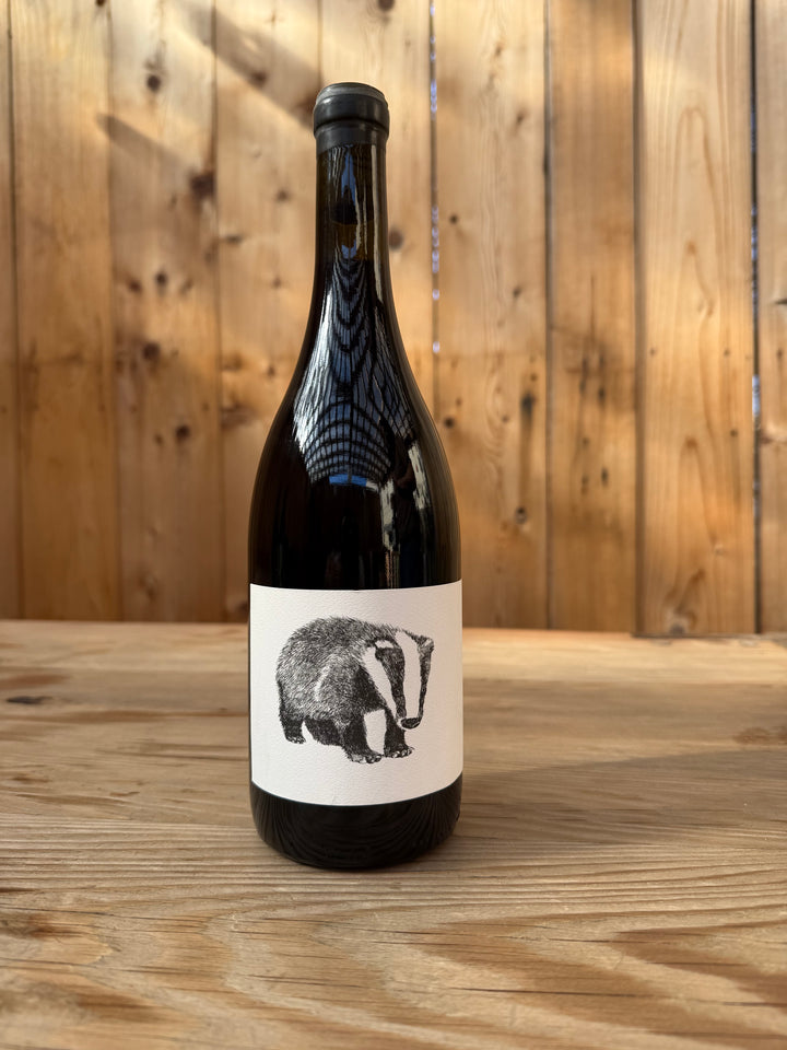 A dark glass bottle of Broc Cellars’ 2018 NERO D’ AVOLA stands on a wooden surface against a wood-paneled background, its white label adorned with a black and white badger illustration. This amphora-aged wine is crafted from Nero d’Avola grapes sourced from Fox Hill Vineyard, distinguished by its striking and artisanal presentation.