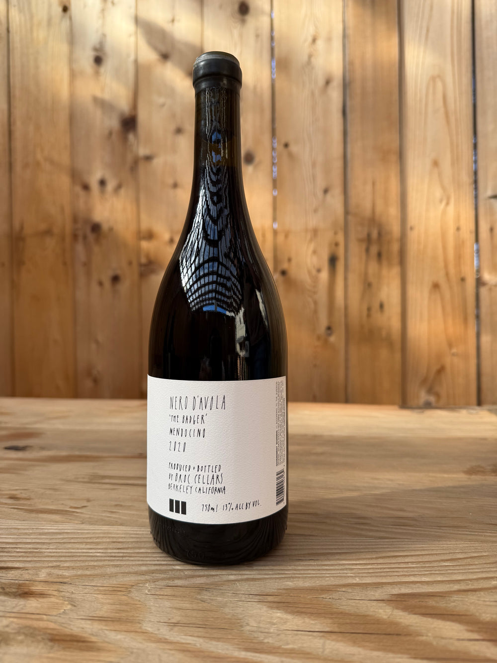 A wine bottle labeled Broc Cellars 2020 Nero d’Avola rests on a wooden surface with a wooden plank wall in the background. The bottle features a white label with black text, highlighting this California wine made from Nero d’Avola grapes.