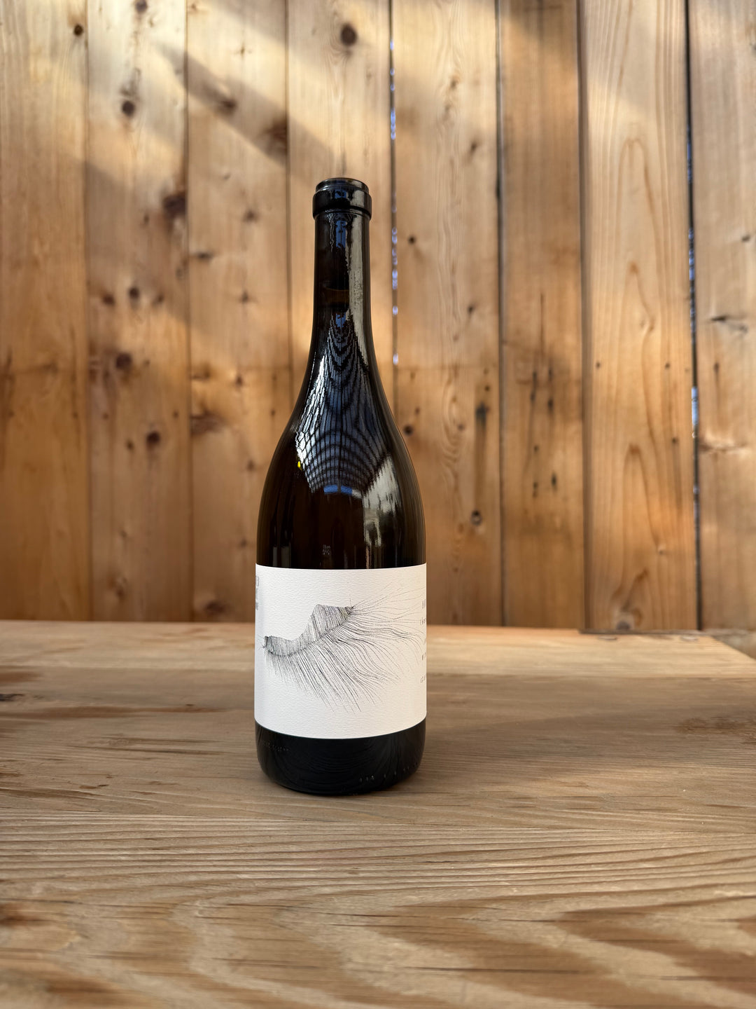 A dark glass bottle with a white label featuring a minimalist black line design sits on a wooden table in front of a wooden plank wall—an elegant presentation for the age-worthy 2022 Michael Mara Chardonnay from Broc Cellars.