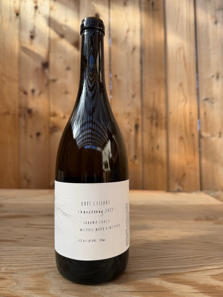 A bottle of wine with a white label sits on a wooden table. The label reads Broc Cellars 2022 Michael Mara Chardonnay, Sonoma Coast, showcasing this distinguished Sonoma Coast Chardonnay. Wooden planks are visible in the background, creating an ideal atmosphere to savor this age-worthy Michael Mara Vineyard selection from Broc Cellars.