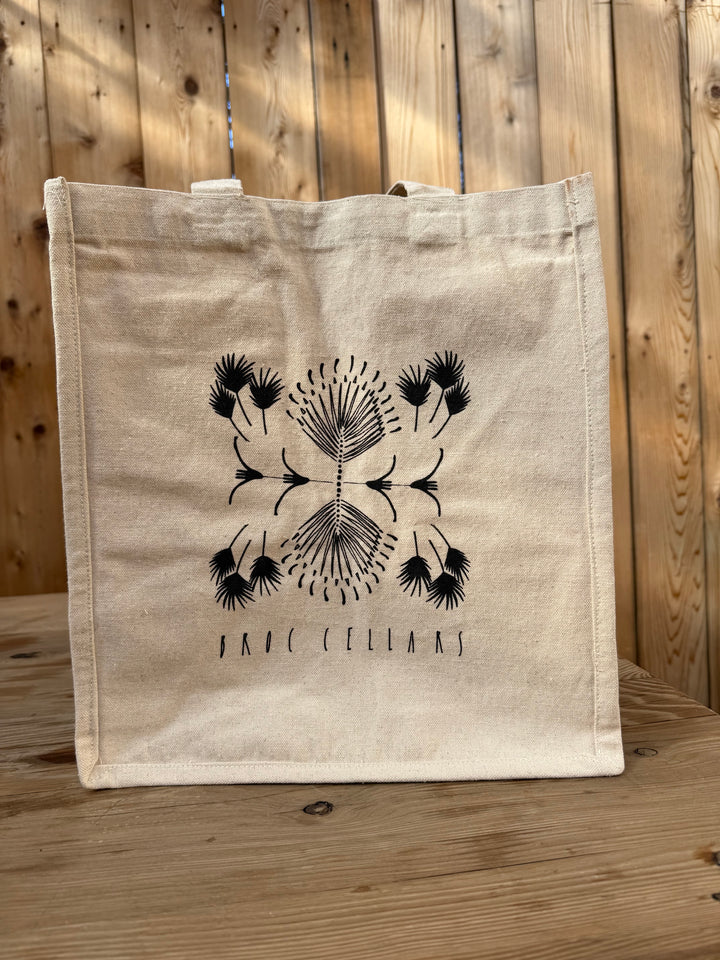 The Broc Cellars Broc 4 Bottle Canvas Bag, a beige canvas tote with handy bottle compartments, displays a black abstract floral design and the “Broc Cellars” logo, featuring artwork by Marta Elise Johansen. It is shown resting on a wooden table in front of a wooden fence.