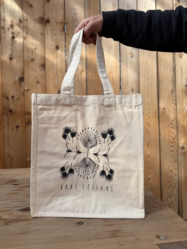 A person holds the Broc Cellars 4 Bottle Canvas Bag, featuring a beige canvas body with a black abstract floral design by Marta Elise Johansen and “BROC CELLARS” printed on it. Designed with convenient compartments for bottles, the bag is displayed on a wooden table against a backdrop of wooden planks.