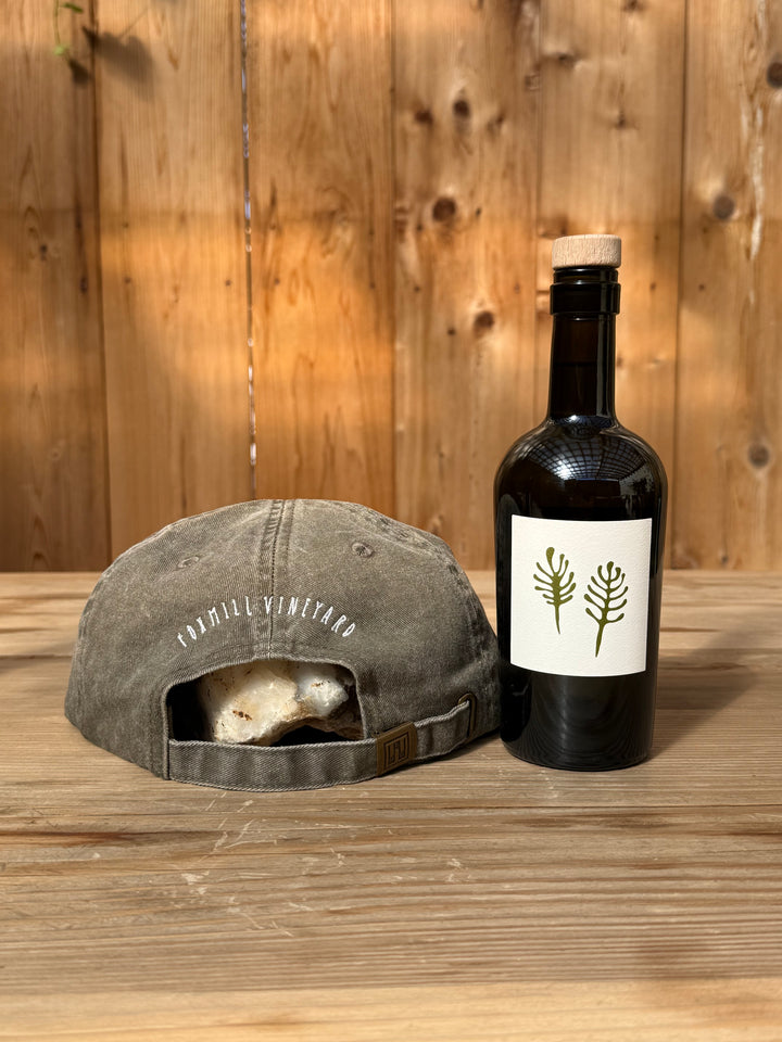 An olive-colored brushed cotton twill Fox Hill Field Hat with white embroidery sits above a rock next to a dark wine bottle with a label depicting two green ferns.