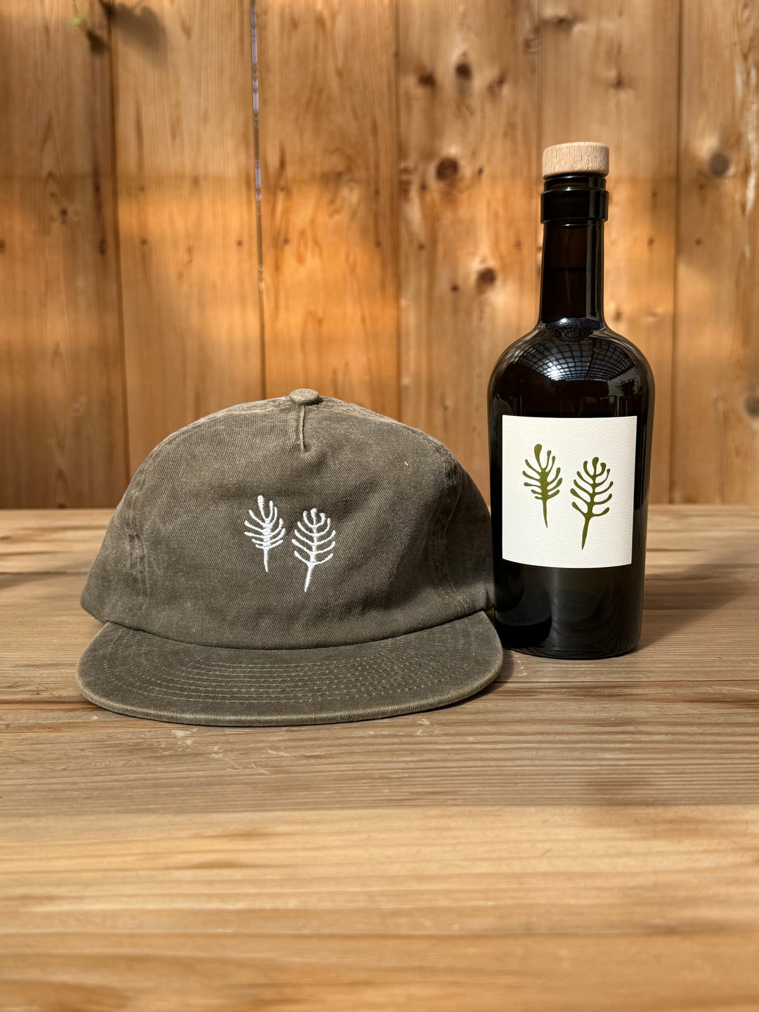 An olive green Fox Hill Field Hat in brushed cotton twill with a white embroidered botanical design sits next to a Broc Cellars wine bottle featuring a matching botanical label.