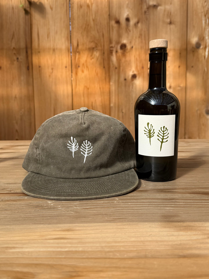 An olive green Fox Hill Field Hat in brushed cotton twill with a white embroidered botanical design sits next to a Broc Cellars wine bottle featuring a matching botanical label.