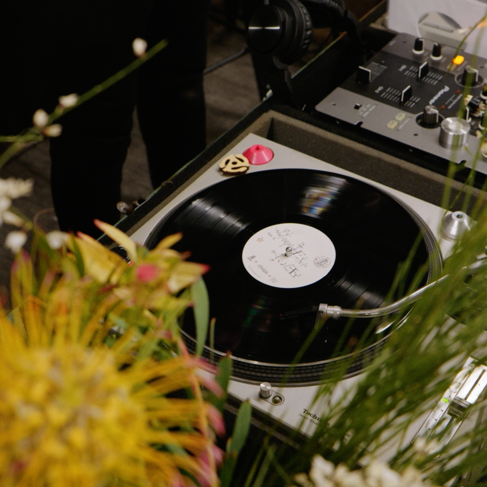 A turntable spins the Broc Cellars “Notes & Tones V.1 + V.2” album duo on vinyl, playing music partly hidden by yellow and white flowers, with audio gear and cables nearby capturing the soulful ambiance.