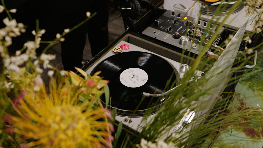 A turntable spins the Broc Cellars “Notes &amp; Tones V.1 + V.2” album duo on vinyl, playing music partly hidden by yellow and white flowers, with audio gear and cables nearby capturing the soulful ambiance.