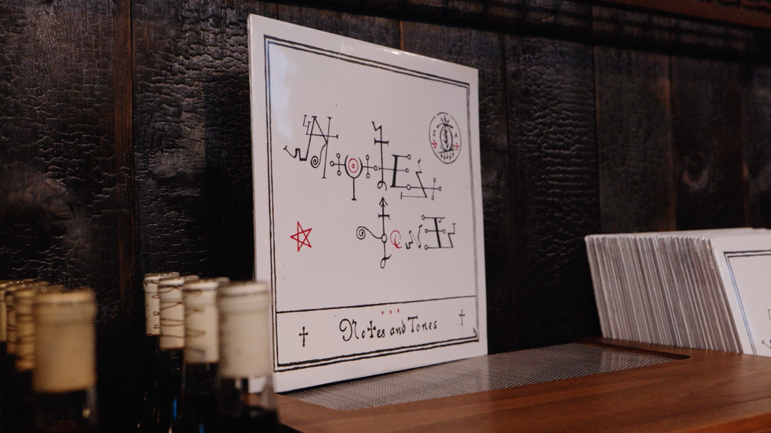 Notes &amp; Tones V.1 - The Album by Broc Cellars, featuring a square white cover with black and red abstract symbols, leans against a dark wooden wall beside bottles and white sleeves, reflecting the brand&