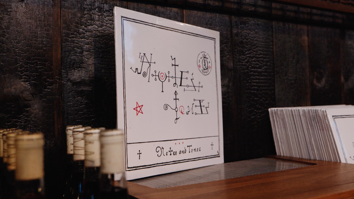 Notes &amp; Tones V.1 - The Album by Broc Cellars, featuring a square white cover with black and red abstract symbols, leans against a dark wooden wall beside bottles and white sleeves, reflecting the brand&