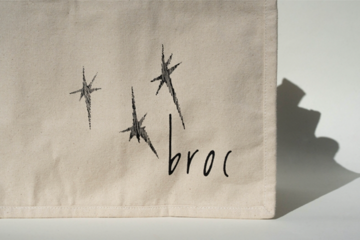 The Broc Bottle Tote by Broc Cellars is a beige canvas tote featuring three hand-drawn black stars and "broc" in lowercase at the bottom right. Includes convenient bottle compartments. Made in America.