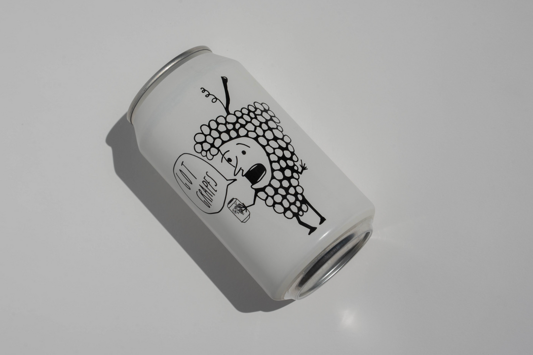 A white Got Grapes Red Can Pack by Broc Cellars rests on its side, showing a black grape bunch with a face holding a drink and saying NOT WINE in a speech bubble—a playful nod to California red wine.