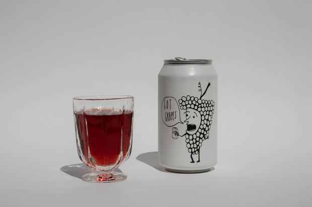 A clear glass of red liquid sits beside a Broc Cellars Got Grapes Red Can, which displays a cartoon grape holding a drink and saying Got Grapes?—coming soon on a plain white background.