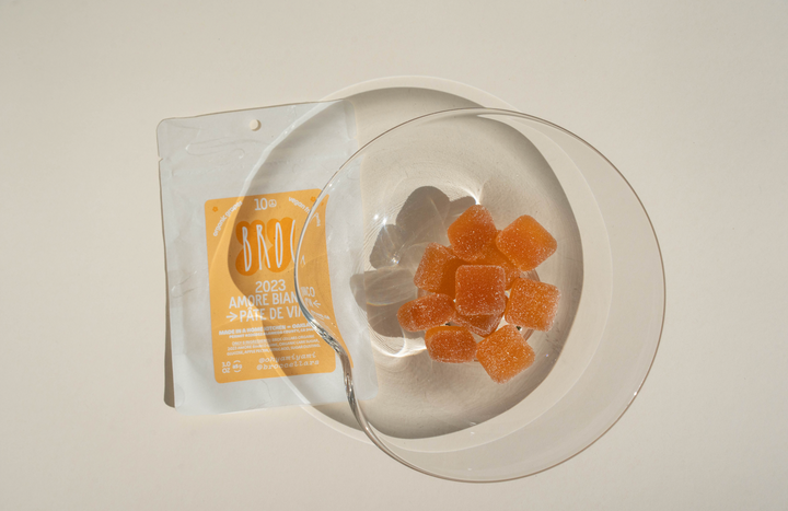 A clear bowl holds orange and translucent Amore Bianco + Yami Yami Pâte de Vin gummy cubes on a white plate. Beside it is Broc Cellars’ silver and orange packet, inspired by skin-contact white wine.
