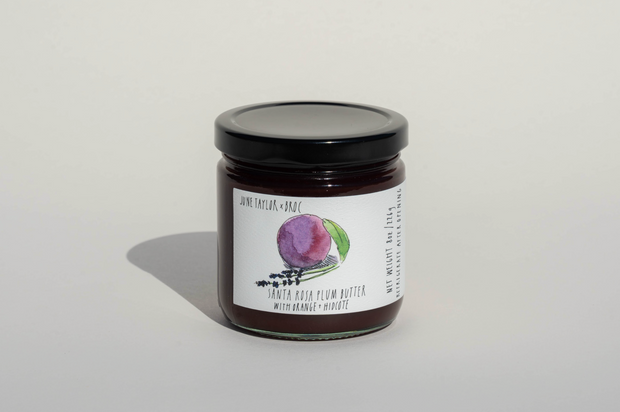 A glass jar with a black lid containing Broc Cellars' June Taylor x Fox Hill Santa Rosa Plum w/ Lavender preserve. The label shows a plum watercolor and reads "Santa Rosa Plum w/ Lavender" with Broc Cellars, June Taylor, and Fox Hill.