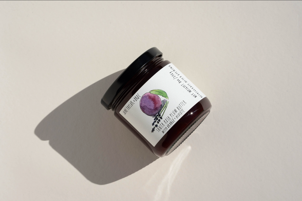 A glass jar with a black lid rests on a beige surface. Its label, adorned with a watercolor plum and leaves, identifies it as Broc Cellars’ June Taylor x Fox Hill Santa Rosa Plum w/ Lavender preserve.