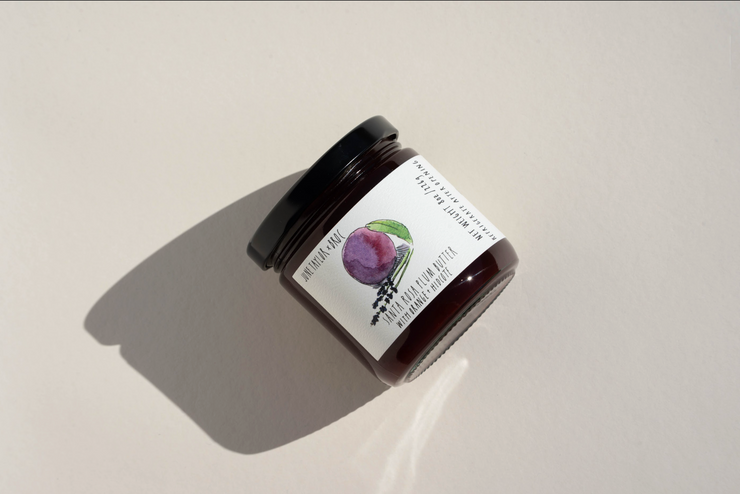 A glass jar with a black lid rests on a beige surface. Its label, adorned with a watercolor plum and leaves, identifies it as Broc Cellars’ June Taylor x Fox Hill Santa Rosa Plum w/ Lavender preserve.