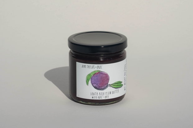 A glass jar with a black lid holds a dark purple spread. The label features a plum and reads “June Taylor x Fox Hill Santa Rosa Plum w/ Bay” by Broc Cellars—a handmade preserve celebrating rich, fruity flavors.