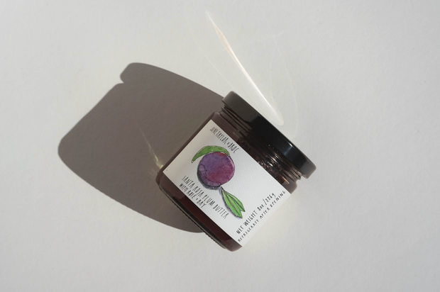 A small glass jar with a black lid rests on a white surface, casting a shadow. Its label shows a purple fruit with green leaves, referencing the handmade June Taylor x Fox Hill Santa Rosa Plum w/ Bay preserve from Broc Cellars. Some text is partly unreadable.