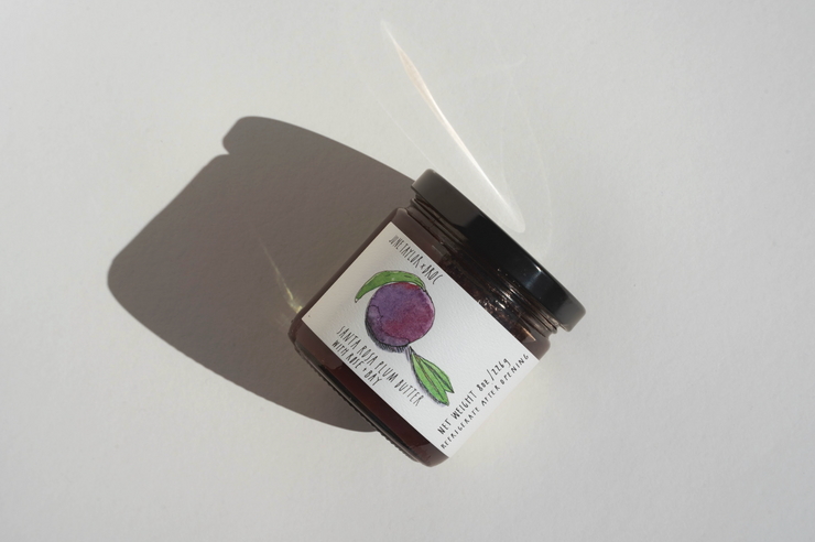 A small glass jar with a black lid rests on a white surface, casting a shadow. Its label shows a purple fruit with green leaves, referencing the handmade June Taylor x Fox Hill Santa Rosa Plum w/ Bay preserve from Broc Cellars. Some text is partly unreadable.