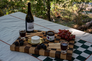On a wooden board atop a table, June Taylor x Fox Hill Santa Rosa Plum w/ Bay by Broc Cellars sits with cheese, crackers, fresh fruit, two glasses of red wine, and a wine bottle outside among sunlit trees.