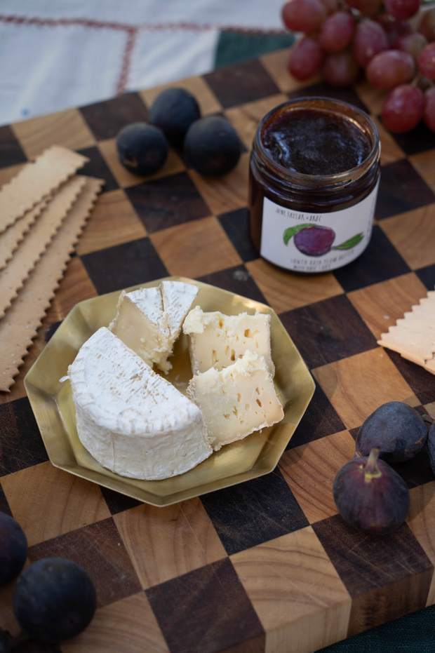A round of soft cheese, partially sliced, sits on a gold plate with crackers, figs, grapes, and a jar of Broc Cellars June Taylor x Fox Hill Santa Rosa Plum w/ Bay fruit preserve on a wooden checkered board.