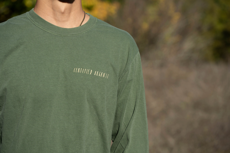 A person wears a Fox Hill Organic Long Sleeve Shirt by Broc Cellars—a green, 100% cotton unisex top with "GRATEFUL AGAINST" in small caps; face hidden, nature background blurred. Comfortable unisex sizing.
