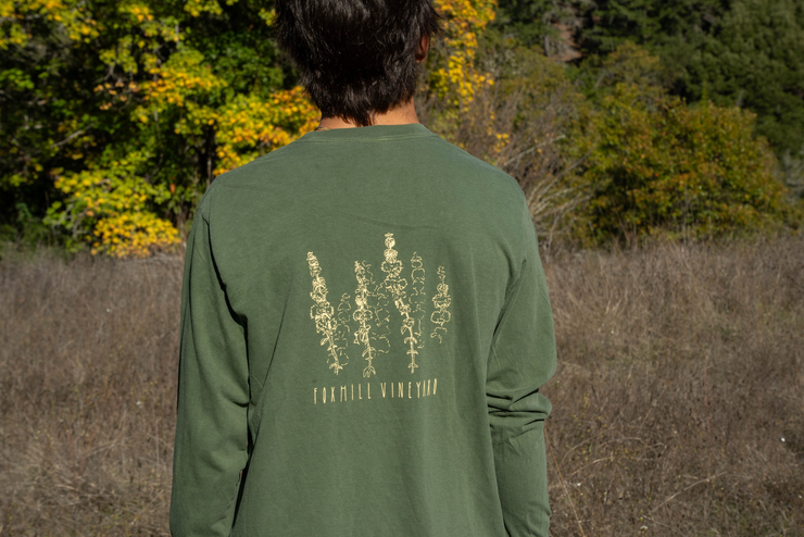 A person with dark hair stands outdoors facing away, wearing a green Fox Hill Organic Long Sleeve Shirt by Broc Cellars, featuring botanical line drawings and trees with dry grass in the background.